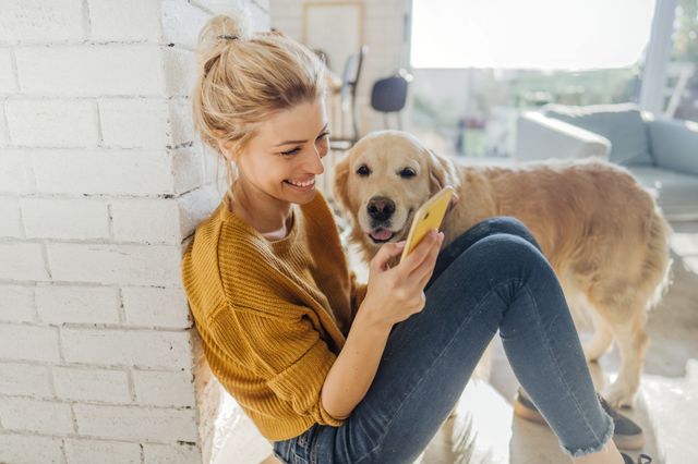 En kvinna sitter på golvet och skrattar medan hon visar något på mobilen för en golden retriever.